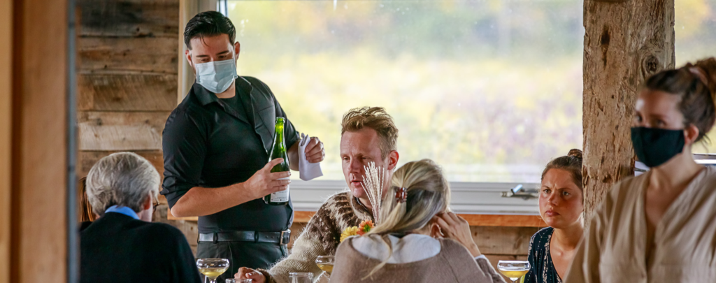 Image of four people seated at in a rustic restaurant. A male server in a mask holding a wine bottle speaks to them. A female server in COVID-19 mask in the foreground.