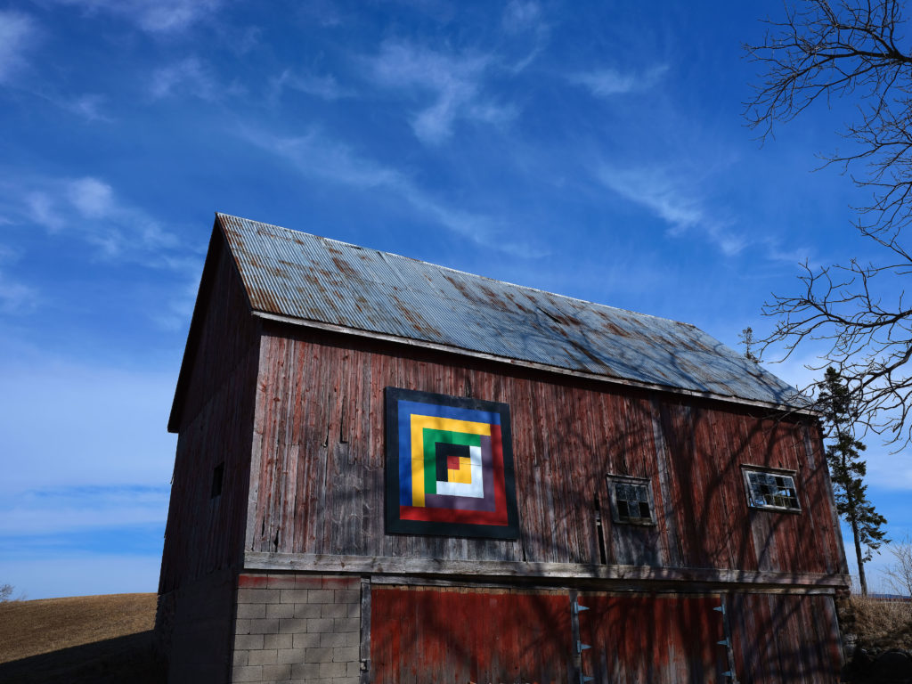 Red barn against a blue sky with a brightly coloured barn quilt above the barn door.