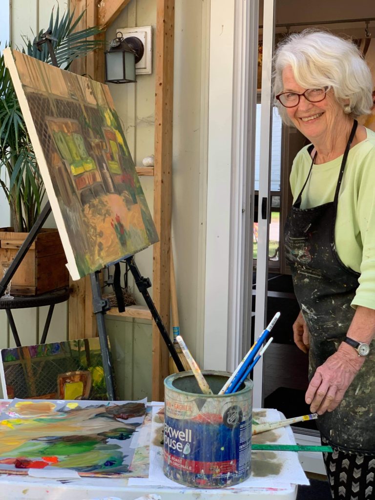 County artist Pamela Carter looking to her left and smiling at the camera wearing a lime green shirt and black painting smock. In front of her is an easel and canvas featuring a work-in-progress painting. In the foreground is a coffee tin filled with paint brushes and a palette of blended paints, the same colours as in the piece on the easel. ⁠