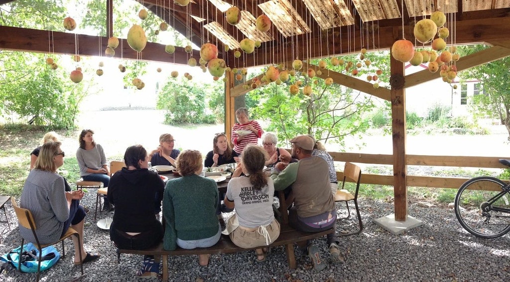 Diners gather around a table under a pergola with hundreds of objects hanging from the ceiling on string above.
